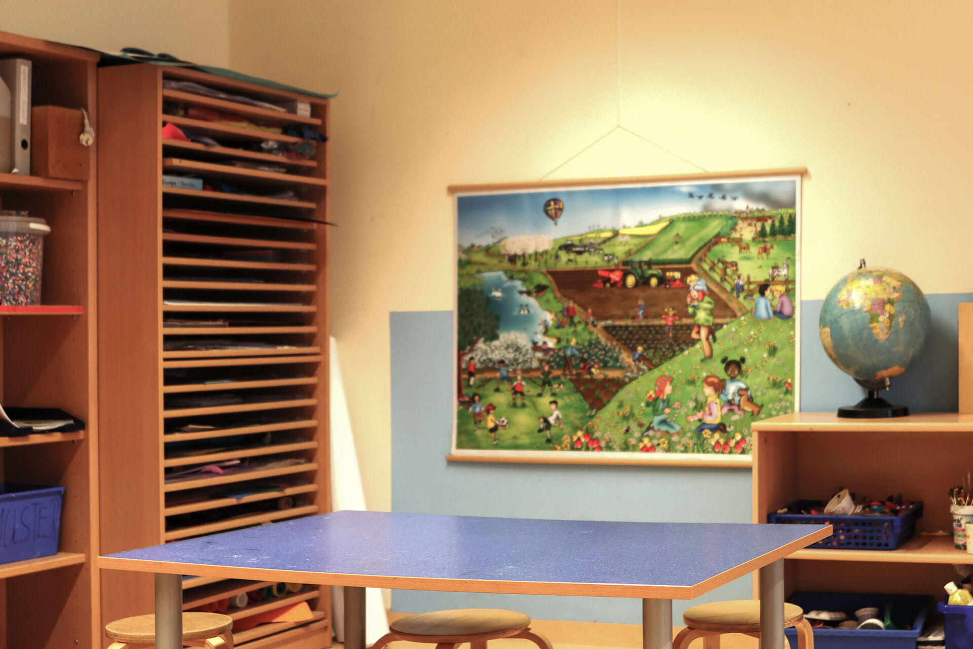 Ein Raum mit hohen Regalen an der linken Wand. Ein Tisch und drei Hocker. Buntes Bild mit spielenden Kindern auf einer Wiese an der Wand.