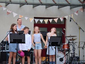 Draußen. Drei Mädchen und ein Junge, dahinter ein junger Mann. Sie stehen auf der Bühne über die bunte Wimpel gespannt sind. Kinder tragen sommerliche Kleidung und freuen sich.