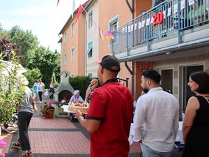 Draußen an einem Haus, Ein Balkon ist mit einer großen roten 20 und AWO Wimpeln geschmückt. Ein Mann mit rotem T-Shirt und Basecape trägt einen Kuchen zum Fest. Neben ihm gehen zwei weitere Personen. Von hinten fotografiert.