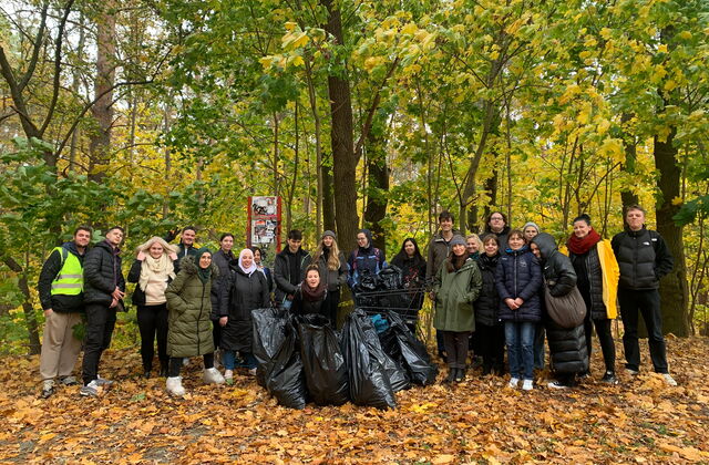 Rund 25 Menschen von Jung bis älter stehen gemeinsam vor einem Wald. Es liegt Laub auf dem Boden aber die Baume haben noch Laub. Vor der Gruppe mehrere gefüllte, schwarze Müllsäcke. Alle blicken stolz und fröhlich zur Kamera.