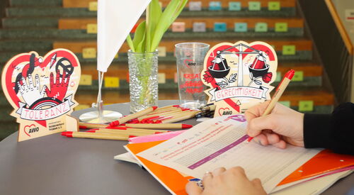 Runder Tisch auf dem eine Unterschriftenliste liegt. Person trägt sich in Liste ein. AWO Kugelschreiber, Becher und Tischaufsteller in Herzform auf dem Tisch.