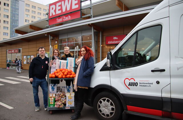Vor einem REWE Markt. Ein weißer Transporter der AWO steht davor. Eine Frau mit langen wilden roten Haaren lehnt am Transporter. 3 weitere Personen schieben einen Rollbehälter beladen mit Lebensmitteln. Alle lächeln in die Kamera.