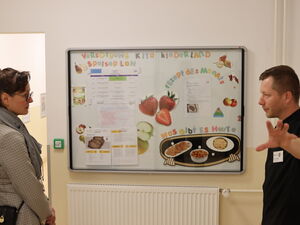 Eine Frau und ein Mann stehen sich gegenüber und sehen sich an. Dazwischen an der Wand ein Schaukasten. Versorgung Kita Kinderland - Speiseplan - Rezept des Monats