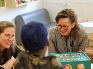 2 Frauen sitzen an einen Tisch mit einer grünen Box. Auf der Bux steht "Wunsch-Essen-Kinder"  Mit dem Rücken zu Kamera ein Kind mit blauem Cappy.