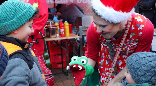 Clownin bückt sich zu zwei Kindern hinunter und lächelt es an. Sie hat eine Handpuppe als Krokodil, das reißt das Maul auf. Clownin hat eine rute Weihnachtmütze auf.