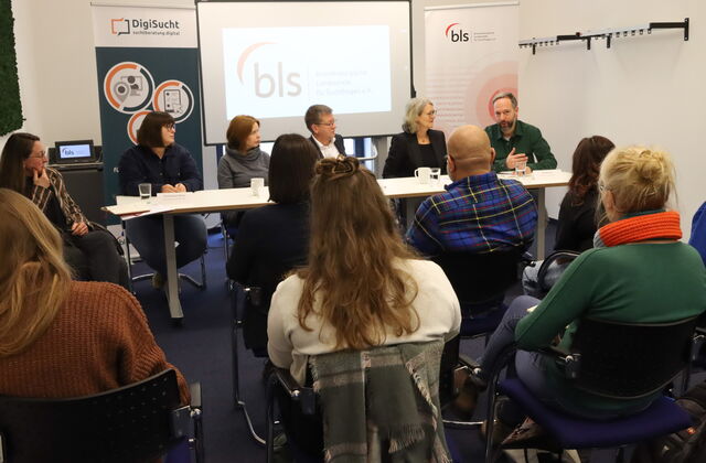 Vier Personen an einem langen Tisch. Drei Frauen und Zwei Männer nehmen an der Diskussion teil.. Zuschauende / Zuhörende sitzen davor.