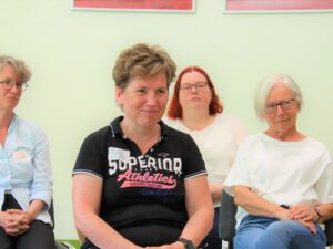 4 Frauen sitzen versetzt auf Stühlen. Frau im Zentrum hat hellbraune Haare, ein schwarzes Poloshirt mit großem Aufdruck vorn.