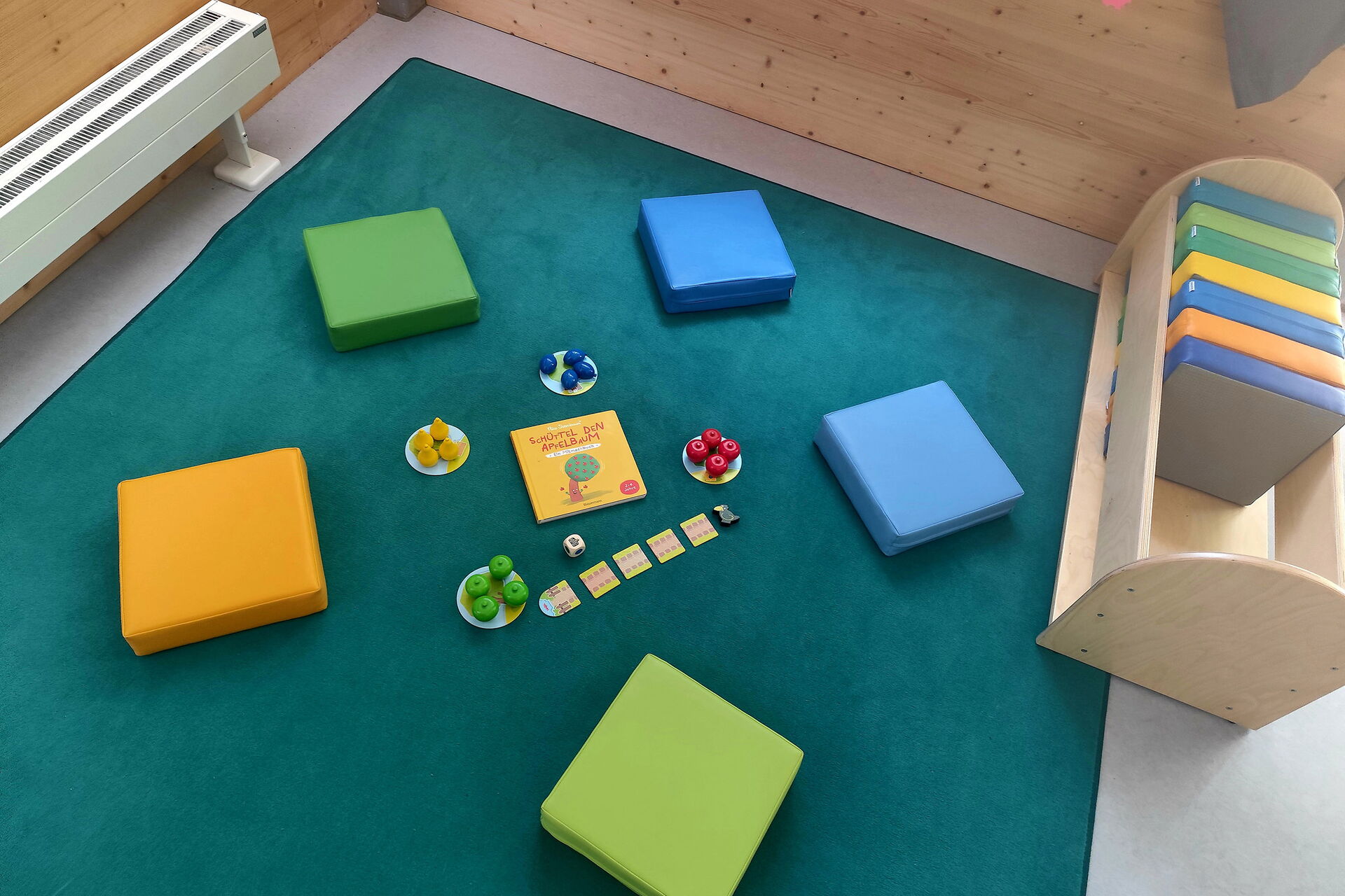 Morgenkreis. 5 farbige Sitzkissen liegen im Kreis. In der Mitte ein Spiel. Weitere Sitzkissen befinden sich in einem kleinen Holzregal.