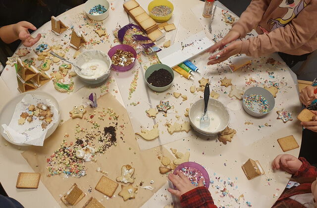 Runder Tisch von Oben. Kinderhände dekorieren Kekse / Plätzchen. Auf dem Tisch sind Butterkekse, Zuckerguss, bunten Zuckerstreuseln, Plätzchen. Alles ist auf dem Tisch verstreut.