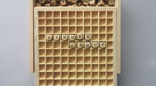 solide Holzbox mit Schiebedeckel beinhaltet Buchstabenwürfel, die in Wörter und Kreuzwortgebilde gelegt werden. Das Wort Kinderarmut ist über zwei Reihen gelegt.