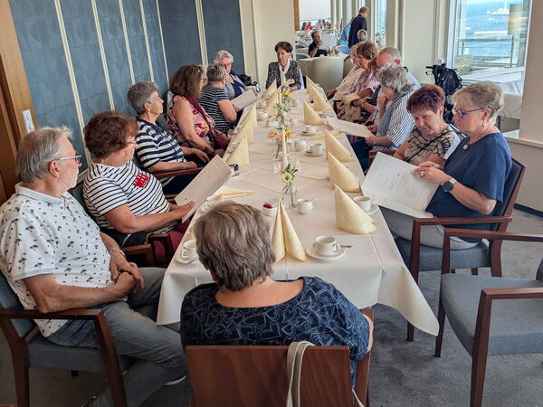 13 Menschen an einer Kaffeetafel in einem großen Raum mit Blick auf Wasser und Schiffe.