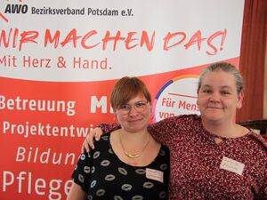 Zwei Frauen stehen vor einem großen AWO Aufsteller. Frau mit braunen Haaren, runder Brille mit goldenen Rahmen, goldene Ohrringe/Creolen und goldener Kette. 2.Frau hat graue Haare die sie im Dutt. Sie trägt eine rote Bluse mit weißen und schwarzen Tupfen.