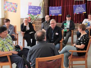 9 Menschen sitzen in einem Kreis. In der Mitte sitzt ein Mann mit blonden kurzen Haaren und Brille mit schwarzem Rahmen.