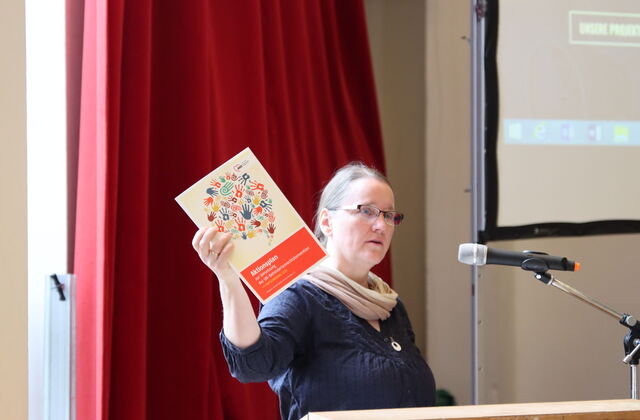 Rosalind Honig bei der Eröffnungsrede des Fachtags. Sie hält dabei ein Exemplar des Aktionplans des AWO Bezirksverbandes mit dem echten Arm und zeigt ihn den Teilnehmer*innen der Tagung