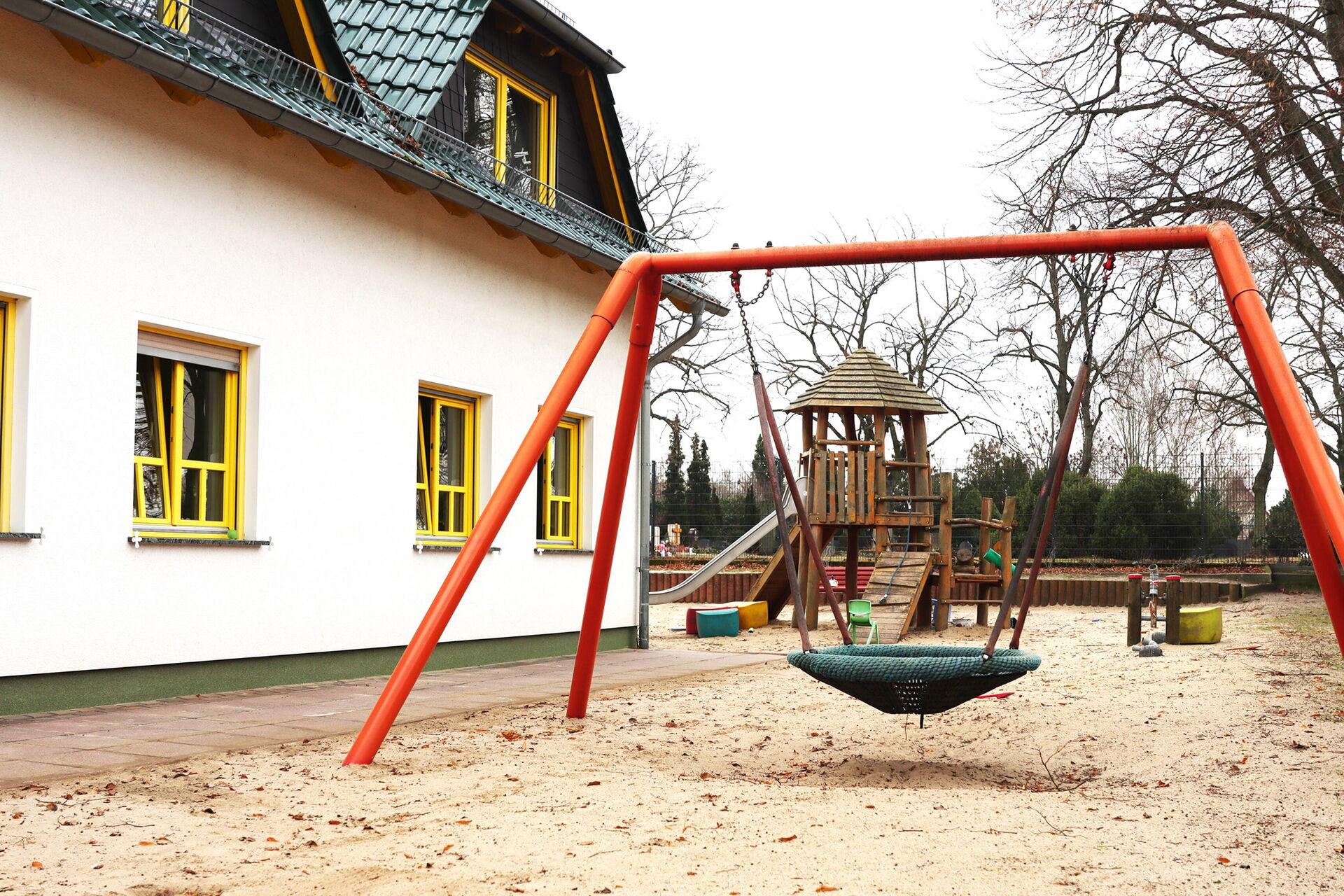 Rechts weißes Haus mit gelben Fensterrahmen und grünem Dach. Spielsplatz mit Vogelnestschaukel und dahinter ein Klettergerüst mit Rutsche.