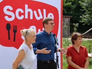 Draußen, Großes Plakat von Schule satt! 3 Personen stehen davor. Ein Mikrofon steht vor einem Redner. Junger Mann mit Brille, kurzen braunen Haaren und blauen Hemd. Links und rechts stehen zwei Frauen.