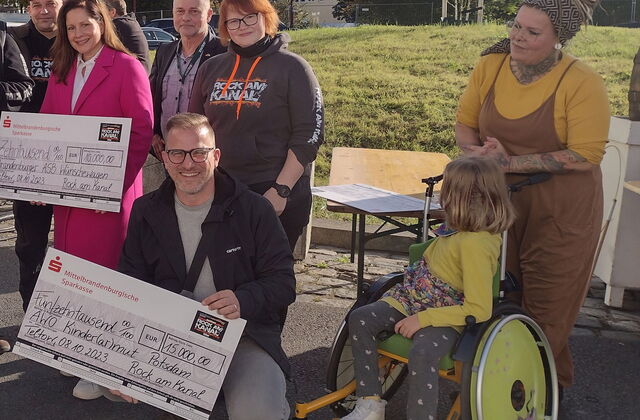 Zwei Spendenschecks werden in die Kamera gehalten. Ein Mann mit Brille, Undercut-Frisur hockt neben Mädchen im gelben Rollstuhl und hält einen Scheck im Wert von 15.000 Euro in der Hand. Dahinter eine Frau im pinken Hosenanzug die den zweiten Scheck hält.
