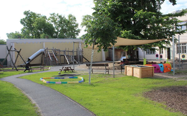 Draußen, großer Spielplatz mit Schaukeln, Klettergerüst mit Rutsche, Sandkasten. Ein großer Baum und ein Sonnensegel spenden Schatten.