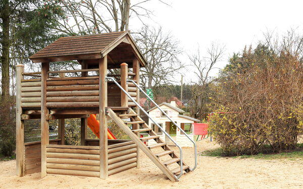 Holzklettergerüst mit Treppe und roter Rutsche im Sand.
