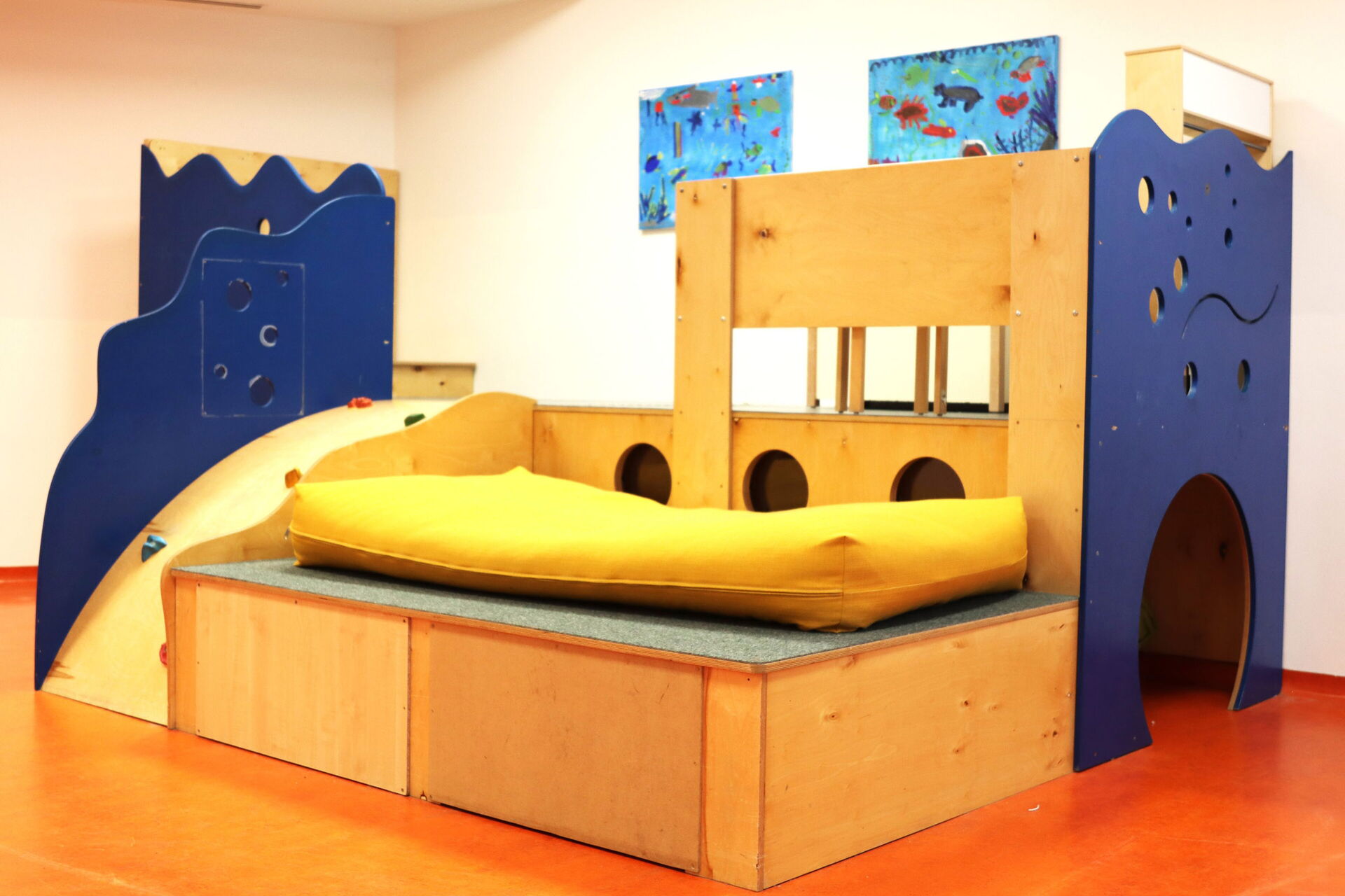 Klettergerüst mit Höhle, Liegefläche mit gelber Matratze, kleiner Rutsche. Es ist aus Holz, einige Elemenze sind blau und Wellenförmig wie das Meer.