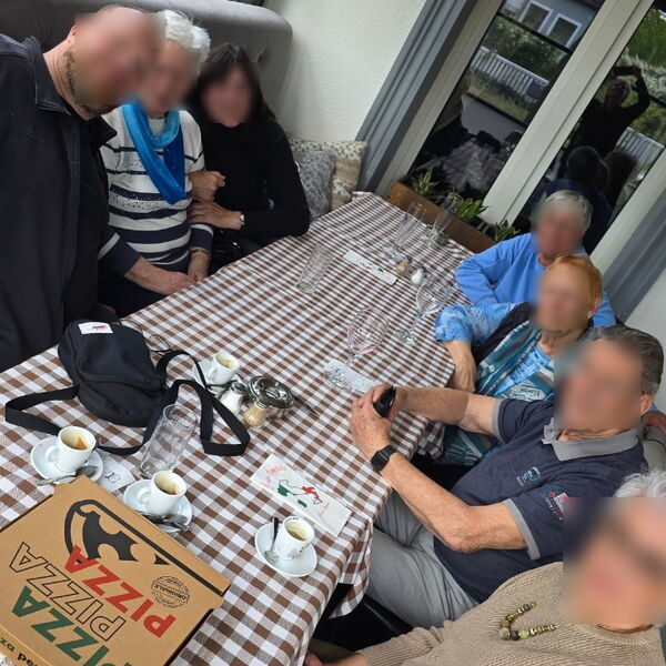 8 Personen an einen Tisch. Auf dem Tisch ein Pizzakarton und Tassen und Gläser.