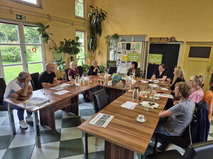 Totale, ganzer Raum mit Tischen in U-Form. 9 Menschen sitzen dort. Gesprächsrunde.