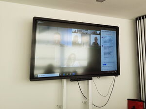 Monitor an der Wand. Ein Online Meeting. Mehrere Menschen sind zugeschaltet.