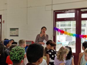 Viele Kinder in einem Raum, die Fenster sind mit Girlanden geschmückt. Eine Frau steht vor den Kindern und hat Papierblätter in der Hand.