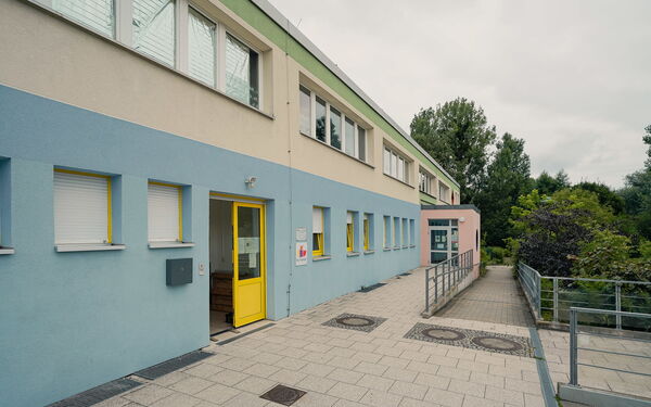 Außen. Gelbe Tür zum Gebäude steht offen. Eine Rampe führt zum Weg hinab. Das Gebäude hat zwei Stockwerke die Fassade ist unten blau, darüber beige und darüber grün.
