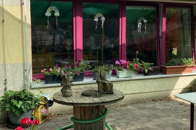 Gebäude mit vielen Fenstern, auf der Fensterbank sind bepflanzte Blumenkästen. Davor steht ein runder Holztisch. An drei der Fenster sind Regenbogenbilder.