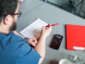 Ein Mann sitzt an einem Tisch, er hat einen roten Kugelschreiber in der Hand und schaut auf ein ausgedrucktes Dokument. Er trägt Brille und ein blaues T-Shift. Ein Handy liegt auf dem Tisch. Blickwinkel über seine Schulter.