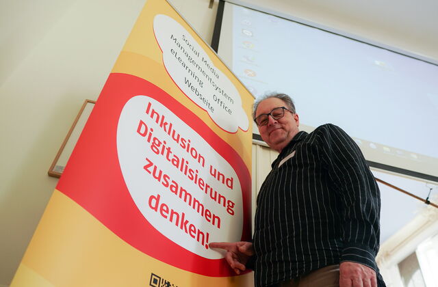 Seminarraum. Von Unten fotografiert. Ein älterer Mann mit Brille, grauen Haaren und dunklem langärmligen Hemd seht vor dem Veranstatungs-Aufsteller. Text: Inklusion und Digitalisierung zusammen denken!