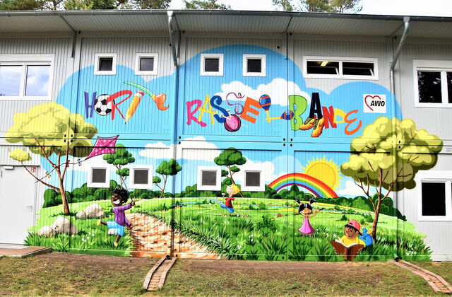 Wandbild an Fassade eines Gebäudes. Bunte Schrift: Hort Rasselbande. Spielende Kinder, Regenbogen, Sonne, Wiese Bäume