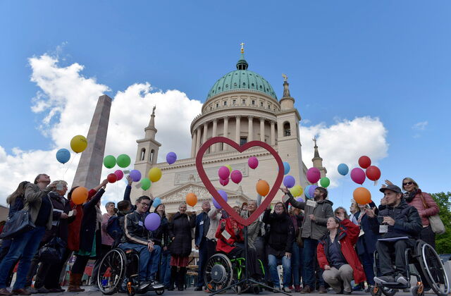 Vor der Nikolaikirche Potsdam, VViele Menschen mit und ohne Behinderung, u.a. drei Rollstuhlfahrer. In der Mitte ist ein großes Herz aufgestellt, und die Menschen lassen bunte Luftballons fliegen.