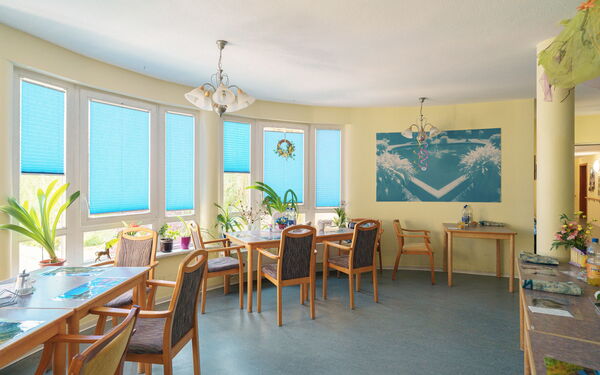kleiner Speisesaal. Freundliche hellgelbe Wände, Tisch mit Stühlen stehen außen wo viele Fenster sind.