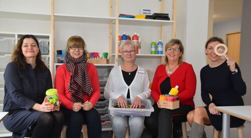 Fünf Frauen sitzen auf Hockern in einem Raum mit Tischen und einem großen Regal.