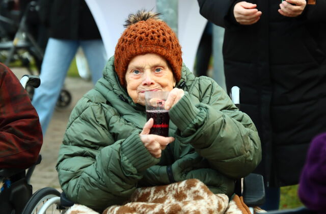 Alte Frau im Rollstuhl. Sie ist dick und warm angezogen mit Decke über den Beinen. Sie hat ein Getränk in den Händen.