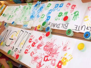 Großer Tisch, viele große Malblätter. Kinder haben ihre Hände in Farbe getunkt um Handabdrücke auf Papier zu bringen, es sind rote, gelbe, blaue, grüne Abdrücke. Text auf einem Blatt: GUTE BILDUNG