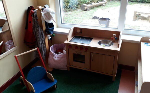 Spielküche aus Holz vor einem Fenster. In der linken Ecke ein Kindergarderobenständer.