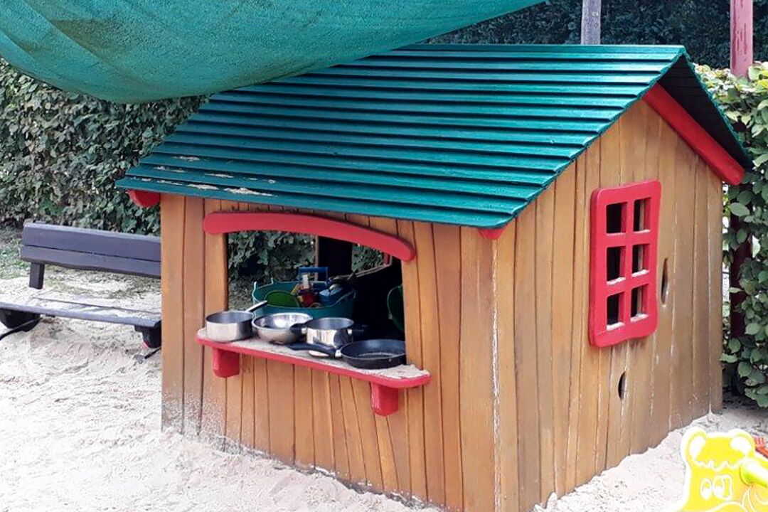 Ein kleines Holzhaus im Garten. Auf dem Fensterbrett Töpfe und Pfannen. Sandkasten davor.