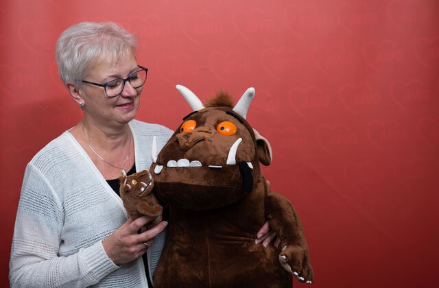 Altere Frau, mit grauen kurzen Haaren, Brille, Ohrstecker und Kette mit heller Strickjacke lächelt ihre zwei Grüffelos an. Ein kleines und ein großes braunes Kuscheltiermonster mit großen Zähnen, Hörnern und orangenen Augen.