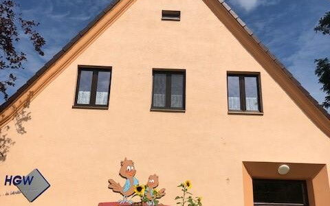 Ein Haus mit Spitzdach und heller Fassade. An dem Haus ist ein Schriftzug:  Kita Spatzenhaus       darüber das Bild von zwei Spatzen.