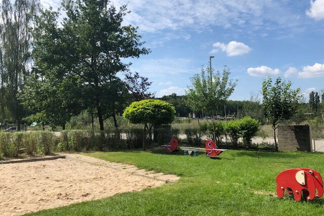 Spielplatz, Sandkasten, auf dem Rasen eine Wippe und ein kleiner Elefant auf dem man reiten kann.