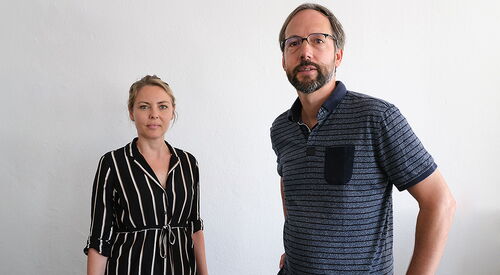 Zwei Personen. Frau und Mann vor einer weißen Wand. Frau ist blond und trägt eine schwarze lange Bluse mit weißen Längsstreifen. Mann mit Brille, Vollbart und blauem Poloshirt.