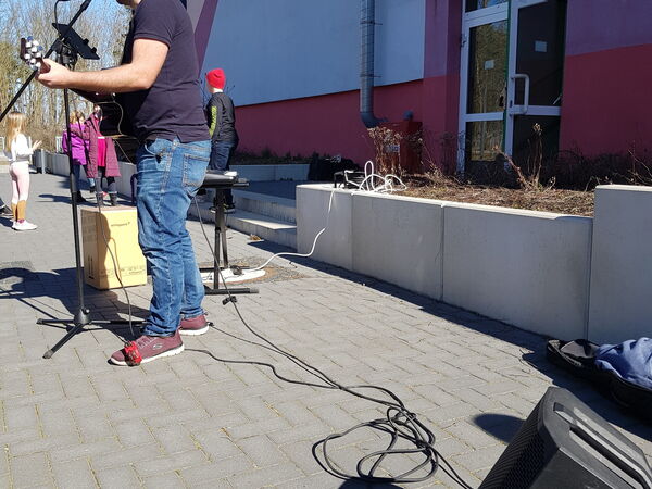 Mann mit Gitarre singt in ein Mikrofon bei einer Außenveranstaltung vor einem Gebäude. Lautsprecher und Kabel auf dem Boden.