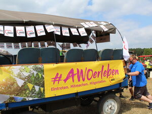 Ein Traktor Anhänger mit #AWO erleben Banner und AWO Wimpeln. Hinten schiebt ihn ein Mann im blauen T-Shirt an.