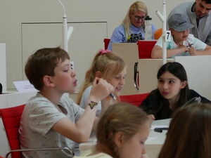Kinder und Erwachsene im Plenarsaal an den Tischen.
