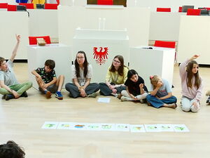 Kinder sitzen in einem großen Kreis auf dem Fußboden des Plenarsaal. In der Mitte liegen Bilder.