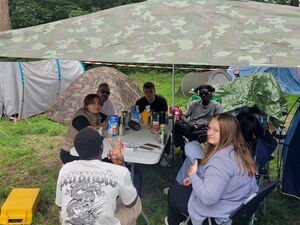 6 junge Menschen auf dem Campingplatz. Sitzen an einem Tisch unter einer Plane in Armee Optik beieinander. Zelte stehen im Hintergrund.