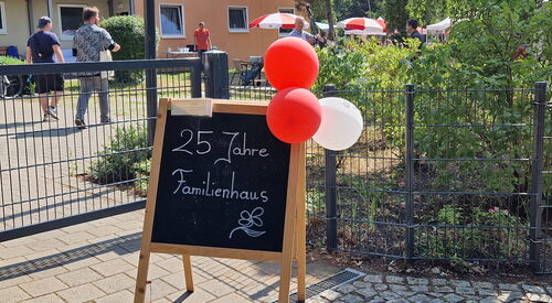 Draußen, Sonnescheint, eine Klapp-Tafel steht am Eingang geschmückt mit roten und weißen Luftballons. Auf der Tafel steht mit weißer Kreide geschrieben: "25 Jahre Familienhaus"